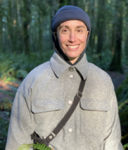 A person with brown eyes and a big smile wearing a navy blue beanie and light grey button up flannel shirt standing in a green forest.