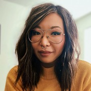 A woman from the shoulders up with long, wavy dark brown hair wearing glasses and jewelry in her nose with full lips in a relaxed smile.