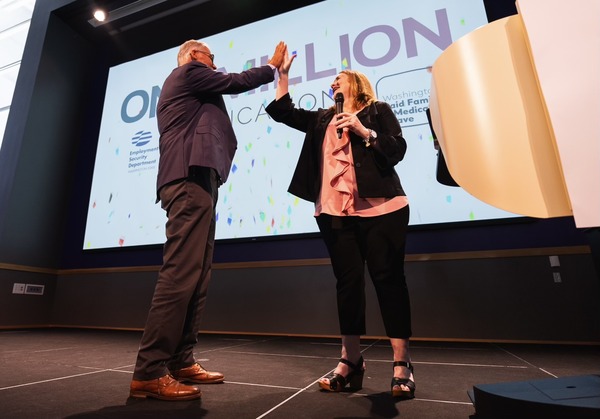 Gov. Inslee high fives a woman on stage in front of a screen that says WA Paid Family and Medical Leave