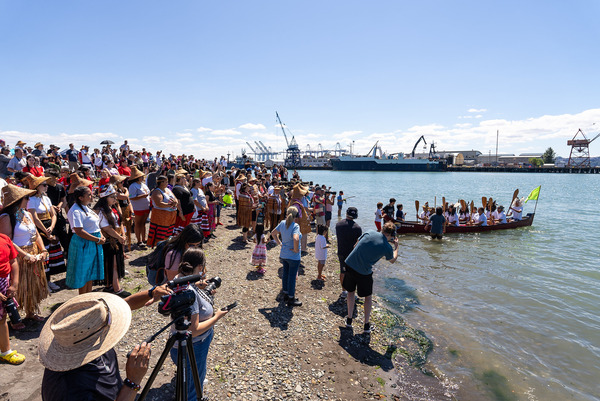 A canoe comes ashore in Tacoma to a large crowd gathered on a beach.