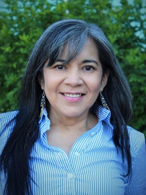 A woman smiles at the camera, with long dark grey hair. She wears a blue and white striped shirt.