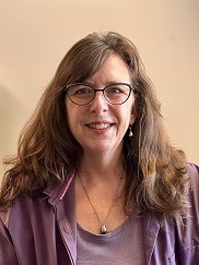 A woman with shoulder length brown hair smiles at the camera. She is wearing glasses.