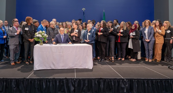 Dozens of people stand behind Gov. Jay Inslee on stage while he signs an executive order