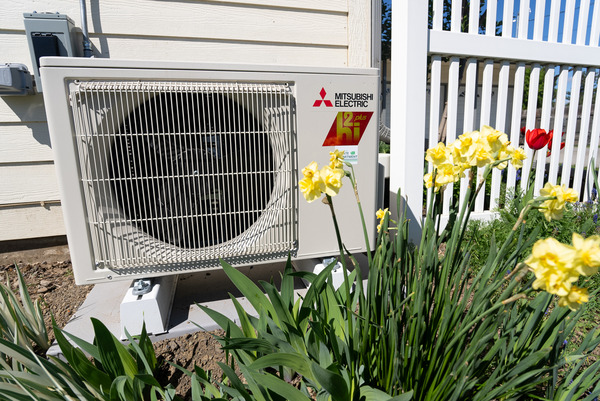 An electric heat pump outside a home near a bed of yellow flowers.