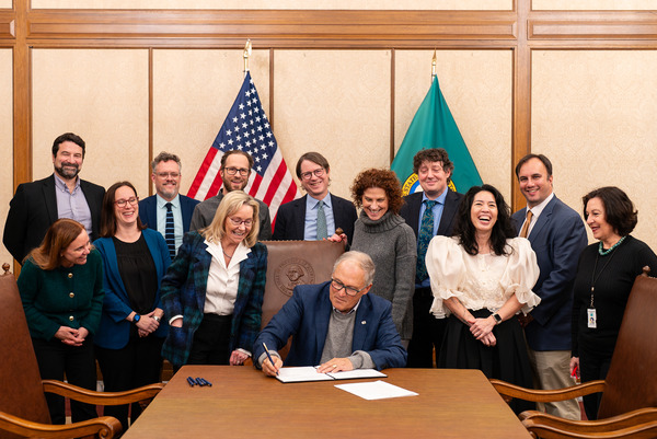 Gov. Jay Inslee signs a document flanked by smiling advisors.