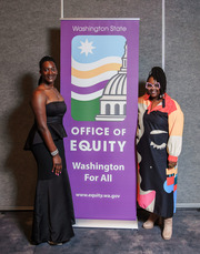 Two Black women smiling standing next to a banner with the Office of Equity Logo & "Washington For All" on it