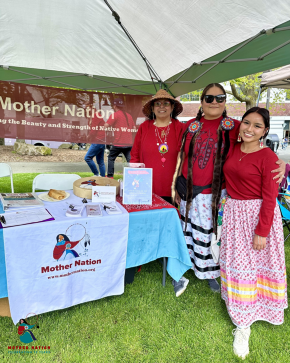 Photo of three women dressed in red, posing in front of a booth for "Mother Nation" and smiling.