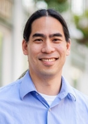 A man smiles, wearing a blue shirt. He has long dark hair parted down the middle and pulled into a low ponytail.