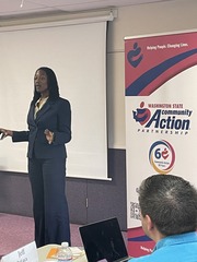 A Black woman speaks in front of a room wearing a suit, standing next to a banner.