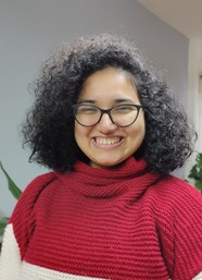 A woman with olive skin, dark curls, a big smile, wearing glasses and a red-cream sweater, framed by green plants on a gray-white background.
