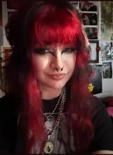 Emma smiling for a selfie, they have red dyed hair and a nose ring in front of a wall of photos