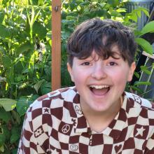 Image of Felix smiling big at the camera. They have brown, slightly curly, short hair. They are wearing a white and brown checkered shirt 
