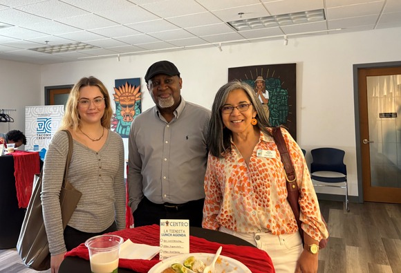Three people stand facing the camera smiling. They are at an event, with food on the table and decorations surrounding them.