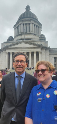 Two people stand smiling outside a large capitol building. One is wearing a suit, while the other wears a shirt with pride stickers.
