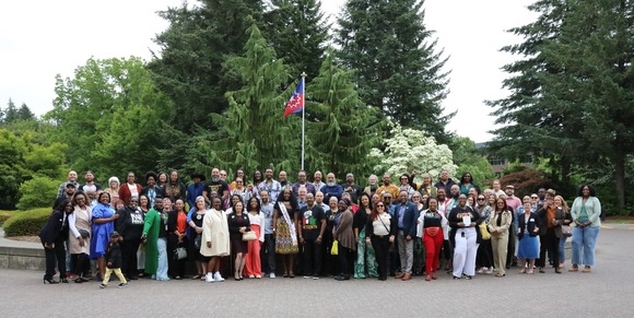 About 100 people stand in front of the Juneteenth flag smiling at the camera. Many of them are dressed up formally in honor of the event.