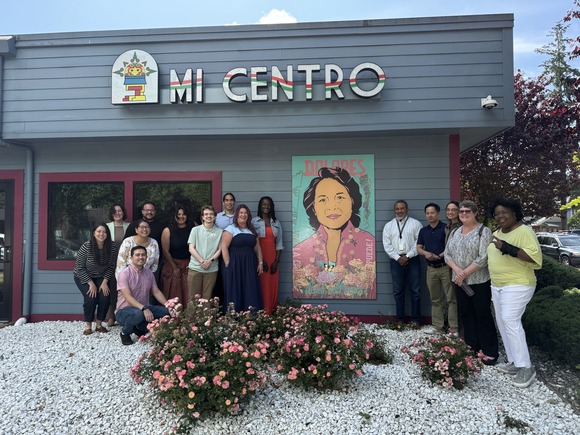 A group of 15 people stand around an image of an art piece of a woman. The sign on the building says Mi Centro.