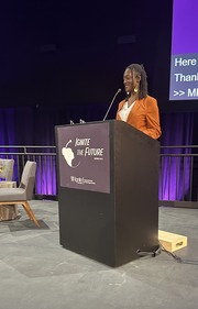 A Black woman stands behind a podium on stage smiling as she speaks. Her audience is not pictured.