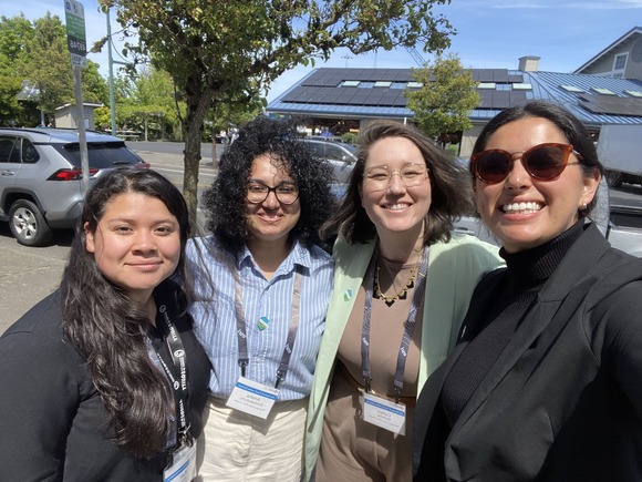 Four people smile at the camera in a parking outside. They are wearing lanyards, like they were just at an event.