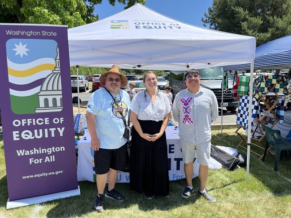 Three people smile at the camera. They are outside in front of an Office of Equity tent & booth.