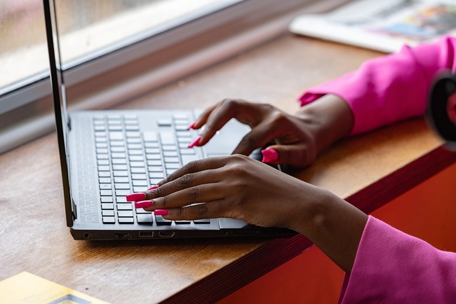 Brown-skin hands with bright pink fingernails typing on a laptop keyboard
