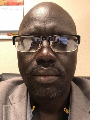 A Black man wearing glasses looks at the camera. He is wearing a grey suit jacket and blue button up.