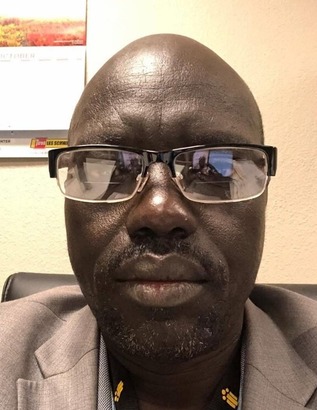 A Black man wearing glasses looks at the camera. He is wearing a grey suit jacket and blue button up.