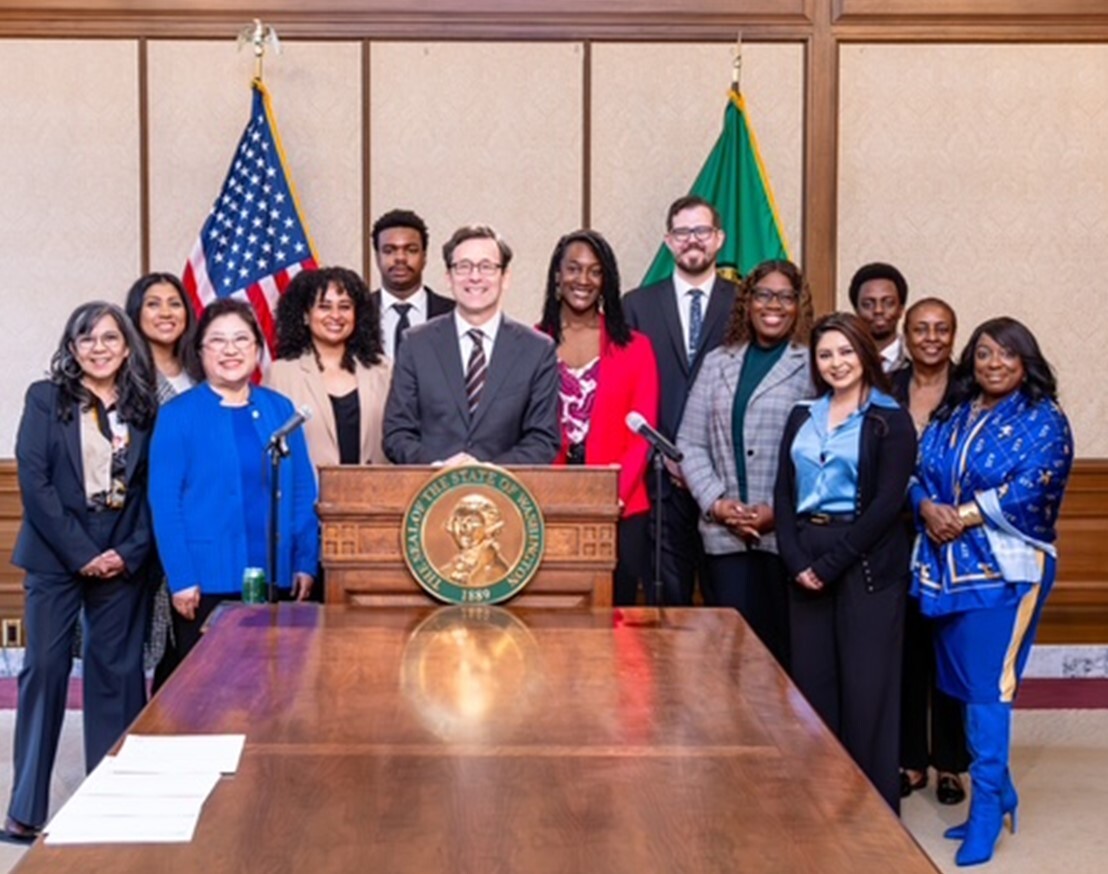 Thirteen people in formal business wear stand smiling at the camera. The man at the center rests his hands on a podium.