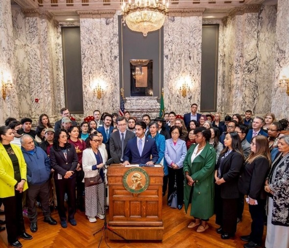 A large group of about 50 people stand around two men in the center at a podium. They are listening to him speak.