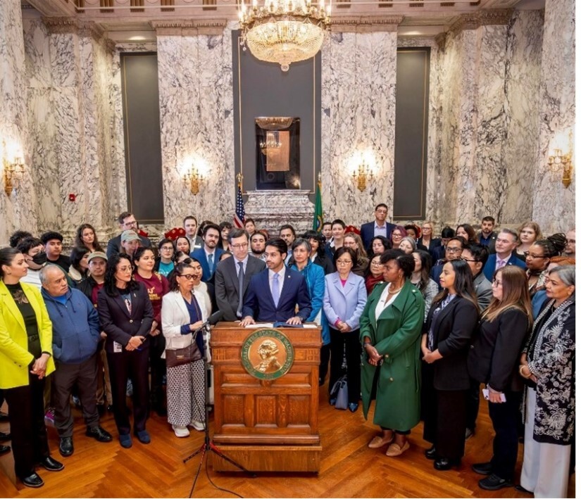 A large group of about 50 people stand around two men in the center at a podium. They are listening to him speak.