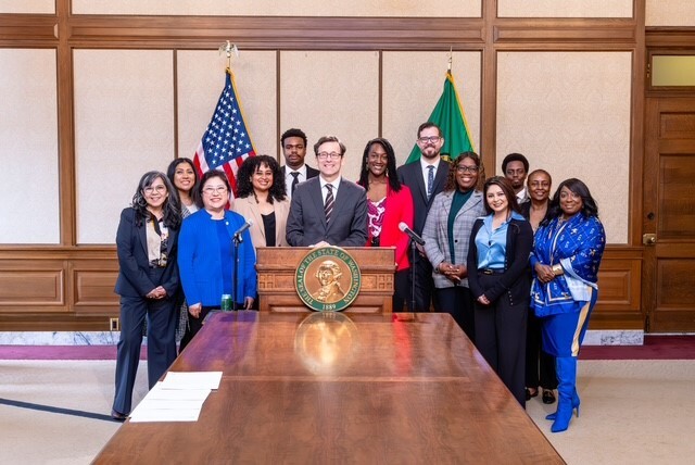 Twelve people stand around the Washington State Governor as he signs a bill.