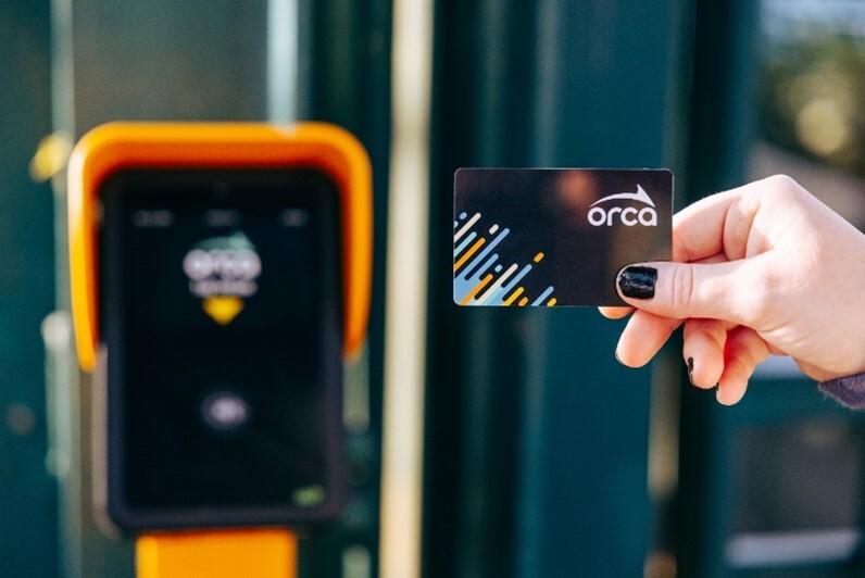A person's hand holding an ORCA transit card up to a yellow electronic reader at a station.