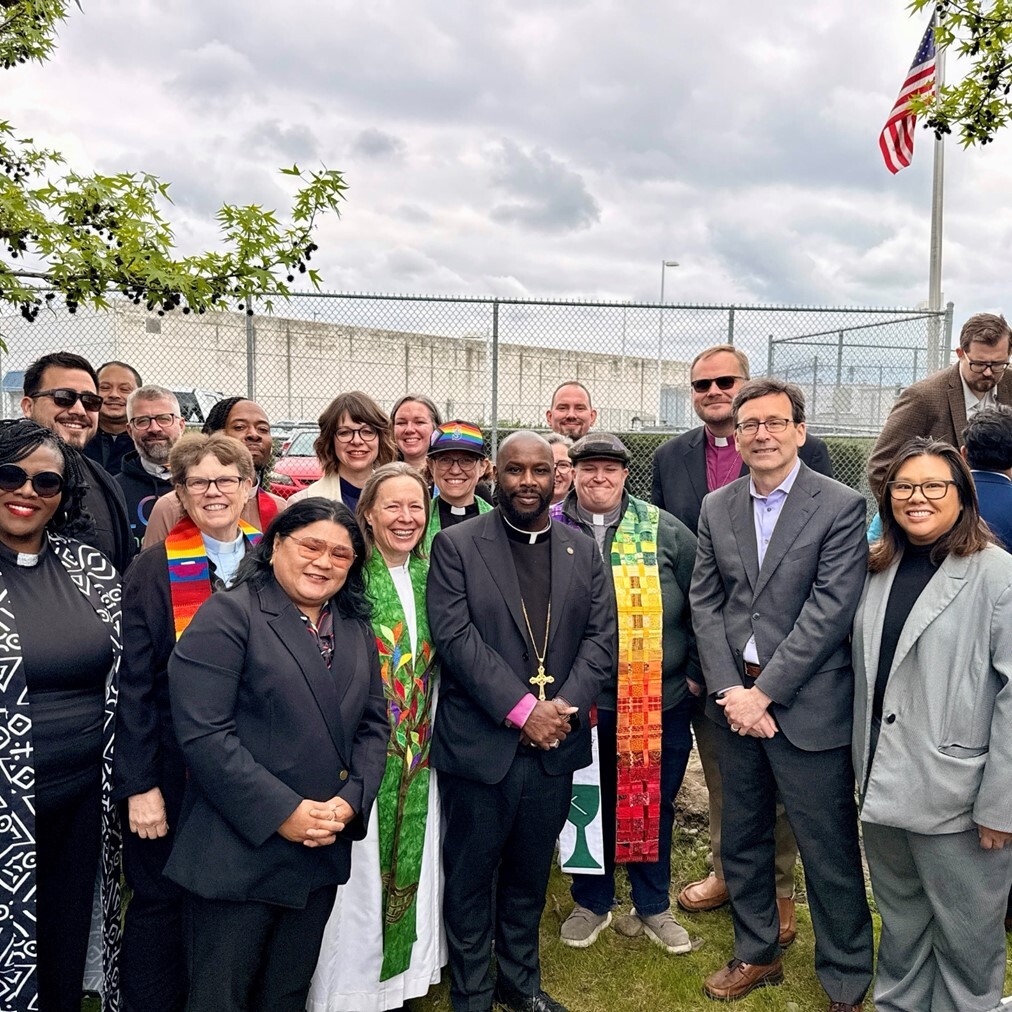 Around 20 people stand with the Washington State Mayor, in front of a large fence.
