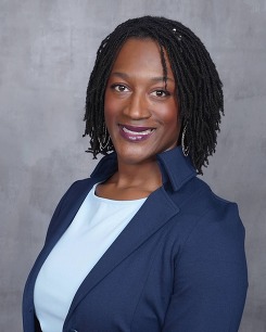 Megan, a Black woman smiles at the camera, her sisterlocks worn loose around her face. She wears a blue button up shirt underneath a black blazer. 