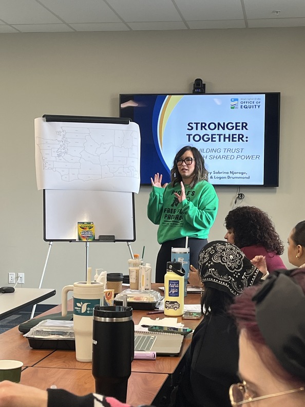 A woman stands at the head of a table leading a meeting. The presentation reads: "Stronger Together: Building Shared Trust with Shared Power."