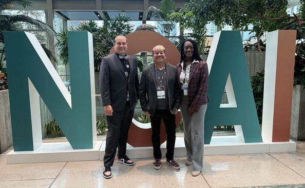 Three people stand next to each other smiling in front of the giant letters "NOAI"