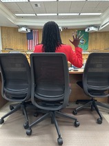 A woman sitting in an office chair with her back to the camera holds her hand up as if she is taking an oath.