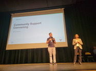 Two people stand on a stage presenting with a slide titles "Community Support Convening"