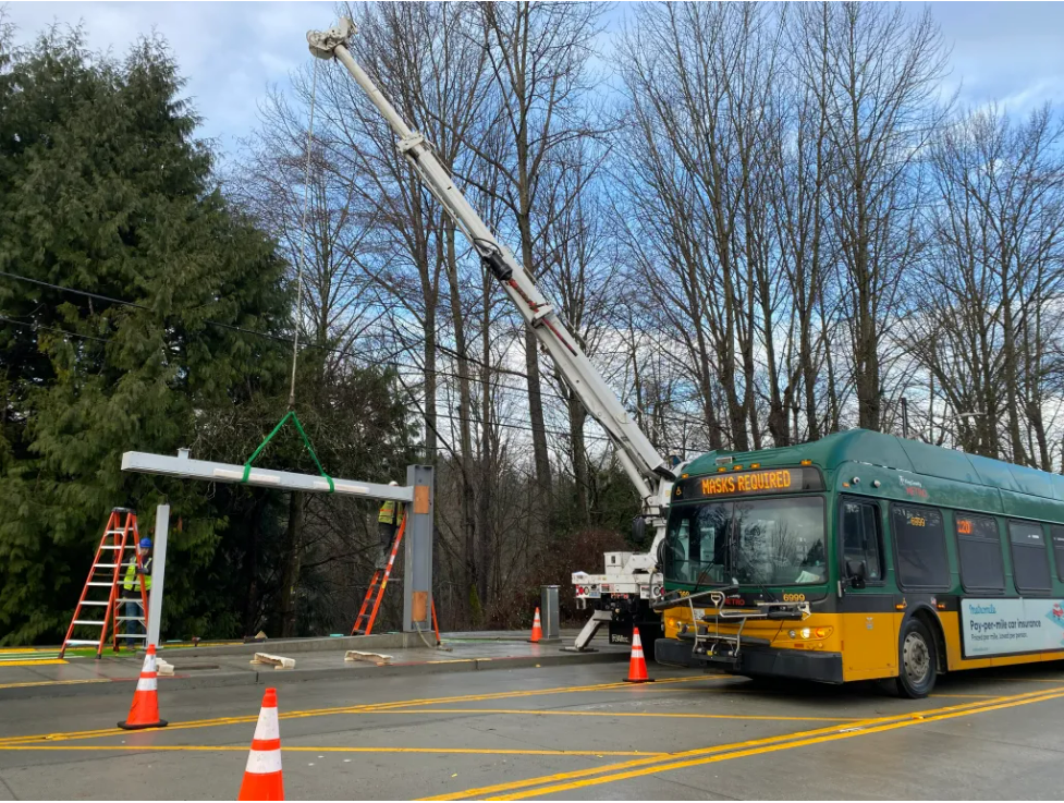 Image showing large equipment installing new RapidRide H Line location. Metro coach to the right, trees and blue sky in background.