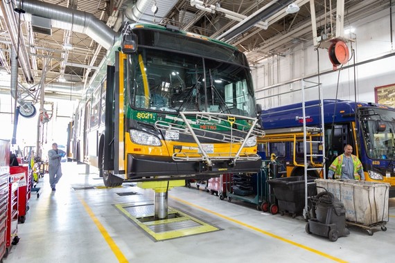 A green and yellow Metro bus is on a large vehicle lift inside a maintenance bay.. Two workers in the facility.