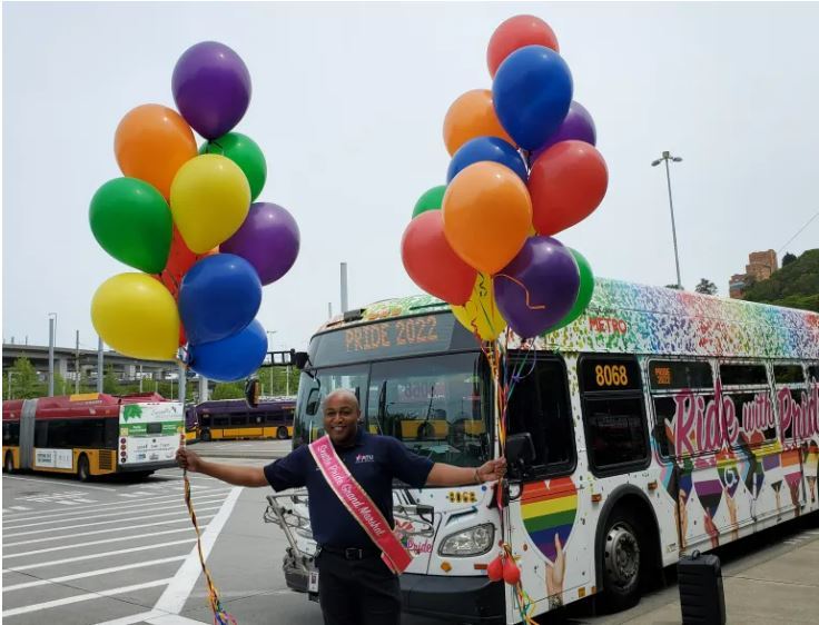 Operator Johnathon Futch named as Grand Marshall, posing with sash and balloons in front of Pride wrapped Metro coach