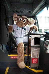 Mariner Moose sits in the operator seat of a Metro bus  and is facing the door of the bus as if to greet boarding passengers. 