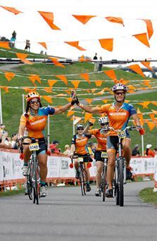 Obliteride cyclists riding on path, while high fiving, beneath orange banners
