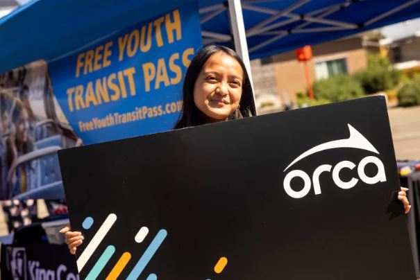 young girl holding an oversized black ORCA card in front of a sign that says free youth transit pass