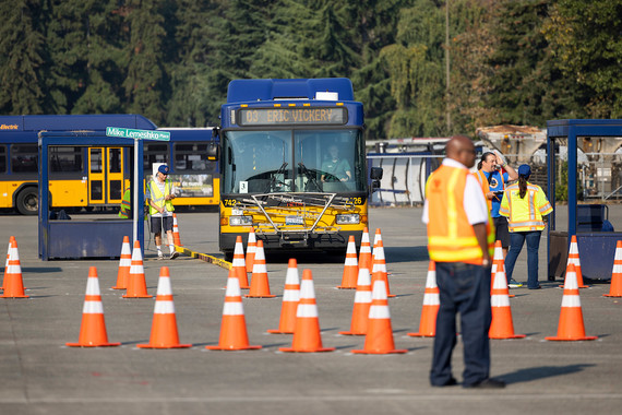 A blue & yellow metro bus is driven on a training facility. bus shelter is set up, in front of it are orange traffic cones marking competition element