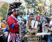 unitedindians.org of a festival with a crowd &two indigenous people talk while wearing cultural dress with headdresses, bells, and other art
