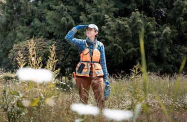 person walking in an orange work vest in a field of tall grass, using hand as visor to block sun from eyes, not looking at the camera