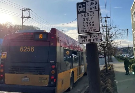 Picture of the back of a RapidRide D line that is pulled over to the side of the road with a bunch of signs and buildings in the background