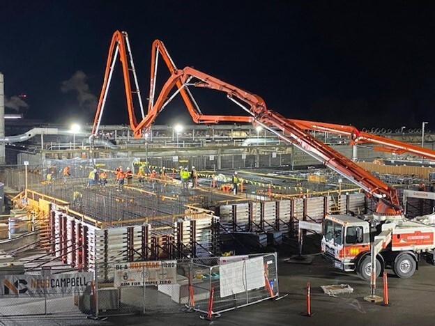 Construction site with lights on at night with concrete trucks using large boom equipment to pour concrete from overhead into foundation forms.