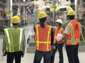 A worker in a hard hat and safety vest points to wastewater treatment equipment and explains to three visitors wearing hard hats and safety vests.