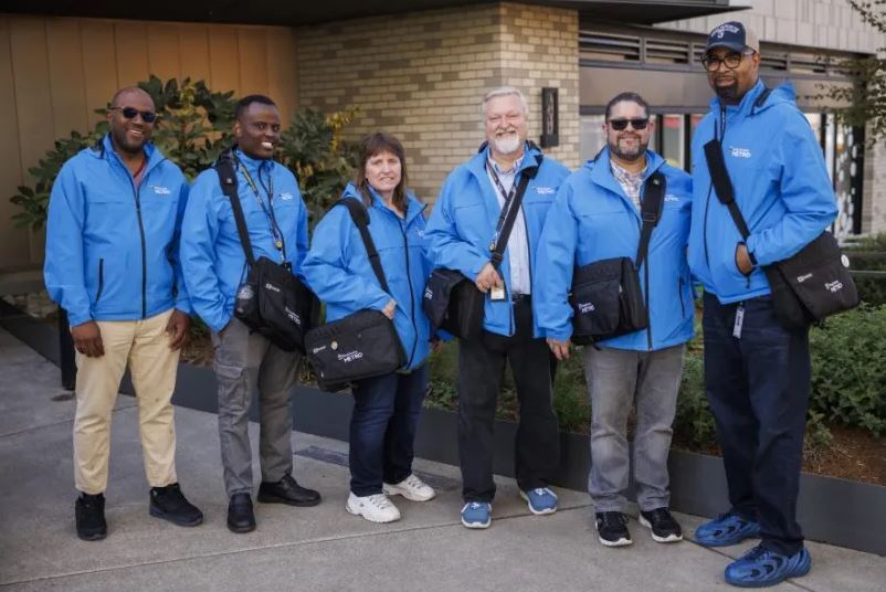 Image of six Metro employees standing in a line smiling while wearing light blue jackets
